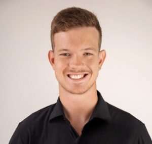 A smiling man with dark, close-cropped hair, wearing a black button-up shirt with the top button unbuttoned.